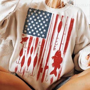 May include: Cream-colored sweatshirt featuring a distressed American flag design with fishing-themed elements. The flag's blue star field is present, with red stripes replaced by fishing rods, hooks, and fish illustrations. A unique patriotic and fishing-themed apparel.