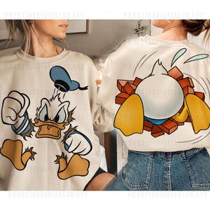 Pode incluir: Camisola de cor creme com um desenho animado do Pato Donald. A frente mostra o Pato Donald com uma expressão zangada, enquanto a parte de trás o retrata a romper uma parede de tijolos. O design inclui detalhes em azul, branco, laranja e amarelo.