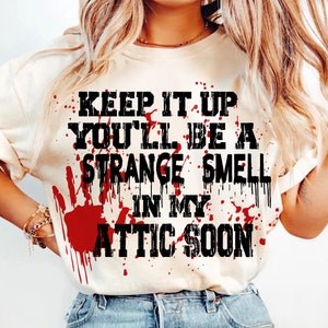 Könnte beinhalten: Cremefarbenes T-Shirt mit dem Text "KEEP IT UP YOU'LL BE A STRANGE SMELL IN MY ATTIC SOON" in Schwarz, mit roten Blutspritzern und einem Handabdruck-Design. Ein lässiges Kleidungsstück.