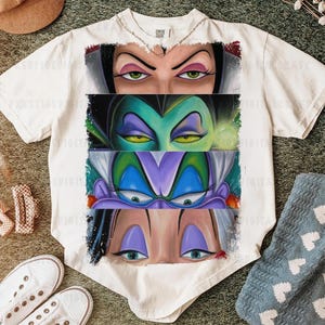 May include: White t-shirt featuring a graphic of four villainous female characters' eyes. The eyes are in a row, each with distinct makeup and expressions. The shirt is on a green surface with shoes and a hat.