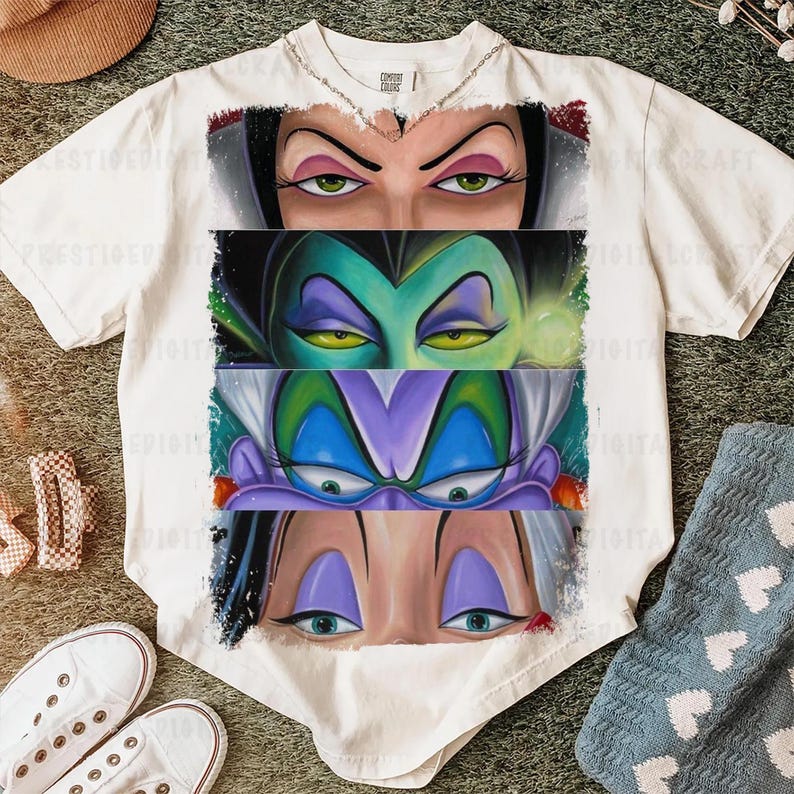 May include: White t-shirt featuring a graphic of four villainous female characters' eyes. The eyes are in a row, each with distinct makeup and expressions. The shirt is on a green surface with shoes and a hat.