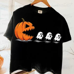 以下が含まれることがあります： ハロウィーンをテーマにしたグラフィックが特徴の黒いTシャツ。 ギザギザの口をしたオレンジ色のジャック・オ・ランタンが、3つの白い幽霊を追いかけています。 このデザインは、古典的なハロウィーンのテーマを遊び心たっぷりに表現しています。