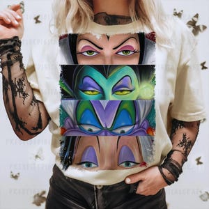 May include: Cream-colored t-shirt featuring a graphic of four sets of cartoon villain eyes. The eyes are in rectangular panels, each with a different color scheme. The shirt is worn by a person with black lace sleeves.