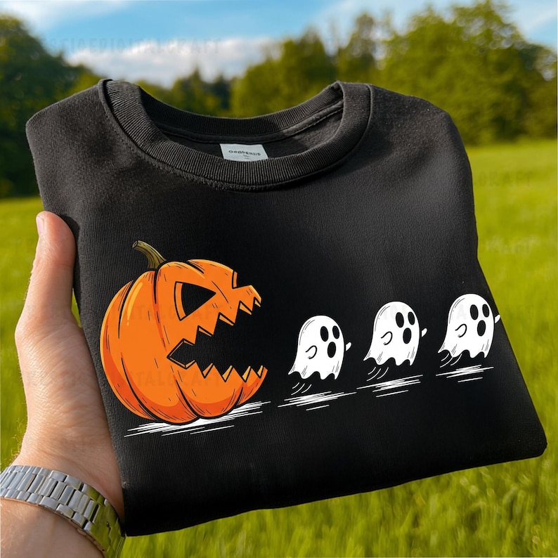 以下が含まれることがあります： ハロウィーンをテーマにしたデザインの黒いスウェットシャツ。大きなオレンジ色のジャック・オ・ランタンが、彫られた口で3つの白い幽霊を追いかけている様子が描かれています。スウェットシャツは折りたたまれており、デザインがはっきりと見えます。