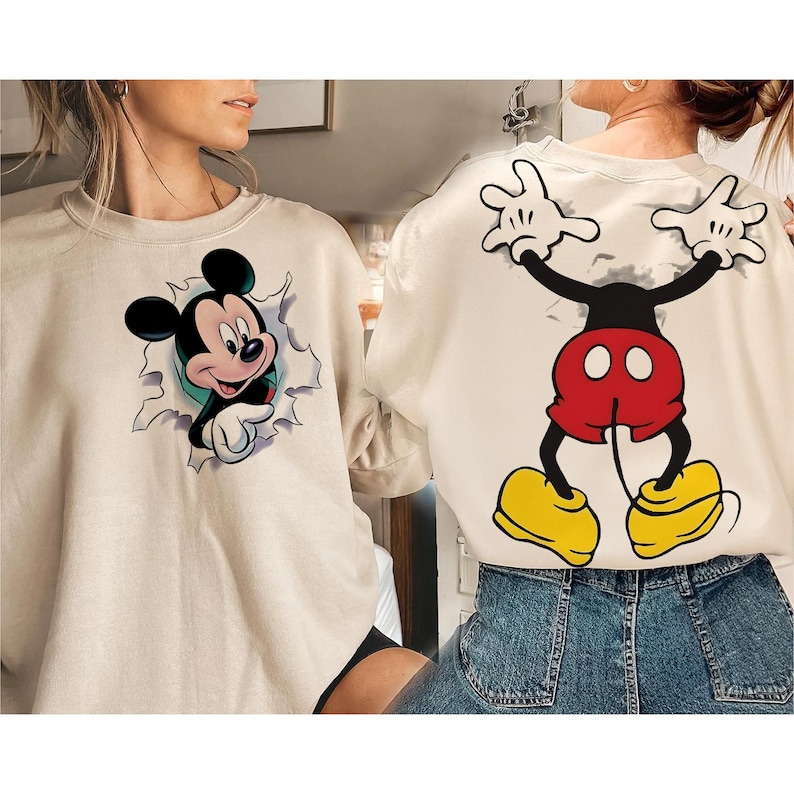 Op de afbeelding: Cr&egrave;mekleurige sweatshirt met een Mickey Mouse-afbeelding. De voorkant toont Mickey die door een muur breekt, terwijl de achterkant Mickey's iconische pose laat zien. Het ontwerp bevat zwart, rood, geel en wit.