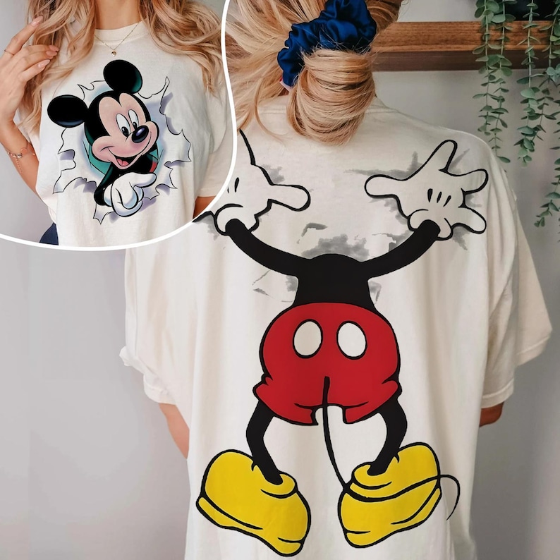 Op de afbeelding: Cr&egrave;mekleurig T-shirt met een Mickey Mouse-afbeelding. De voorkant toont Mickey die door een gescheurd ontwerp breekt, terwijl de achterkant een volledig beeld van Mickey in rode shorts en gele schoenen laat zien. Een leuke, casual top.