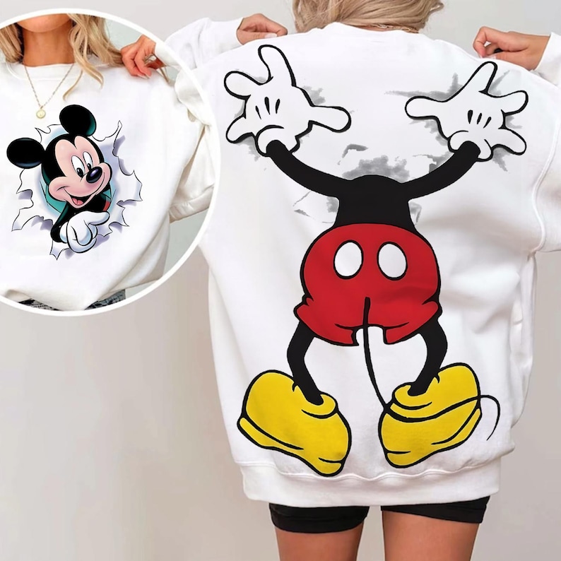 Op de afbeelding: Witte sweatshirt met een Mickey Mouse-ontwerp. De voorkant toont Mickey die uit een gescheurd effect komt, terwijl de achterkant een full-body cartoonontwerp laat zien met een rode korte broek, gele schoenen en witte handschoenen.