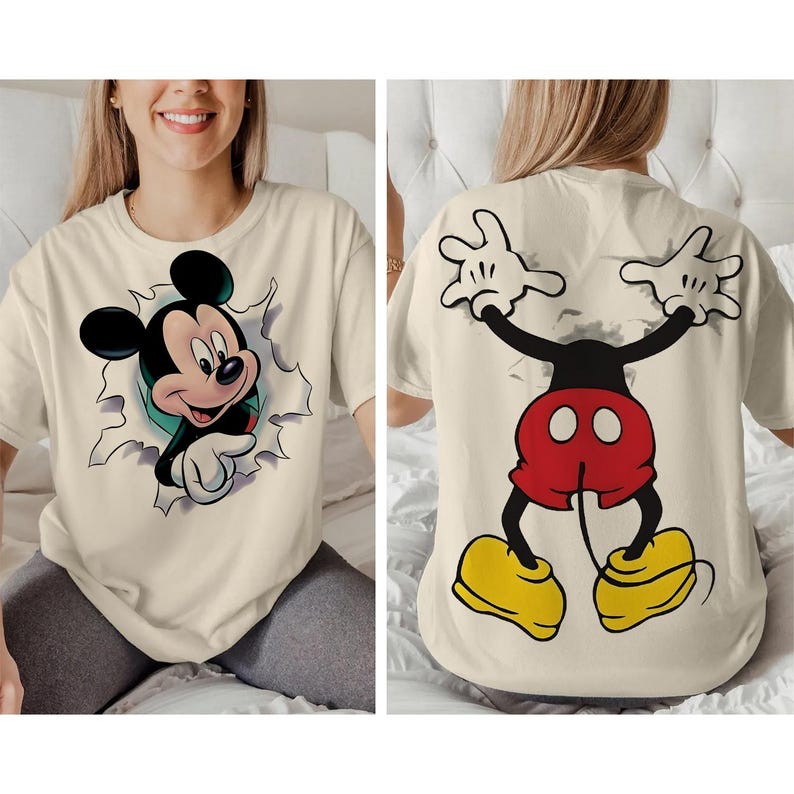 Op de afbeelding: Cr&egrave;mekleurig T-shirt met een Mickey Mouse-afbeelding. De voorkant van het shirt toont Mickey die door een gescheurd muurontwerp breekt. De achterkant toont Mickey's armen, korte broek en schoenen.