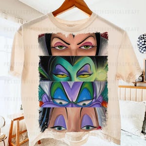May include: Cream-colored t-shirt featuring a graphic of four sets of cartoon villain eyes. The eyes are in shades of green, purple, and black, with detailed eyelashes and eyebrows. The shirt is hanging on a wooden hanger.