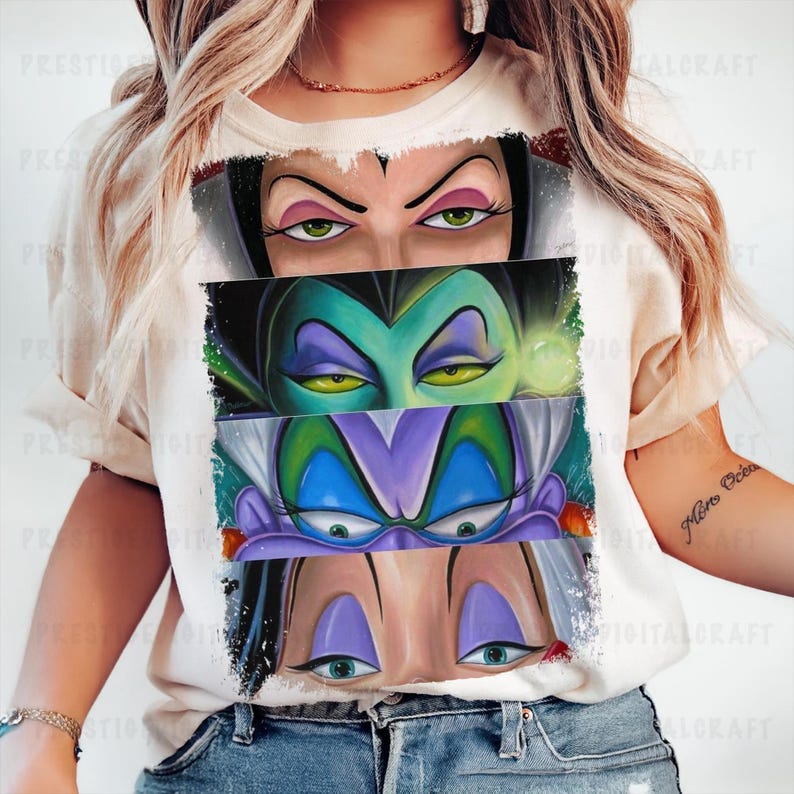 May include: A cream-colored t-shirt featuring a graphic of three sets of cartoon villain eyes. The top set has black hair and pink eyeshadow, the middle set has green skin and yellow eyes, and the bottom set has purple hair and blue eyes.
