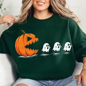 以下が含まれることがあります： ハロウィーンをテーマにしたデザインの濃い緑色のスウェットシャツ。彫刻された顔を持つオレンジ色のカボチャが、3つの白い幽霊を追いかけています。このデザインは、古典的なビデオゲームのユーモラスな解釈です。このスウェットシャツは、快適でカジュアルな衣服です。