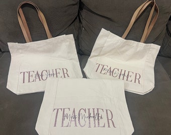 May include: Three off-white canvas tote bags with brown handles. Each bag has the word "TEACHER" printed in a glittery, mauve font, along with a name. The bags are arranged on a dark gray surface.