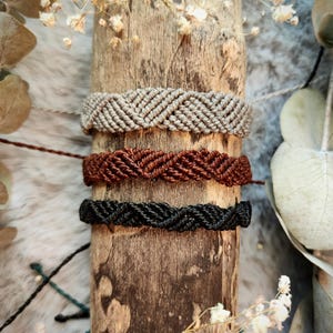 Può includere: Tre braccialetti intrecciati in diversi colori: grigio, marrone e nero. I braccialetti sono impilati su un pezzo di legno.