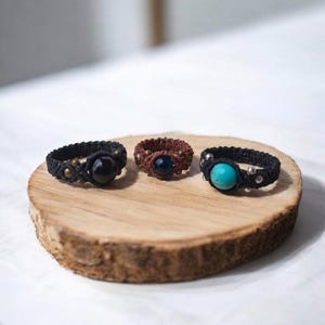 Könnte beinhalten: Drei Makramee-Ringe mit zentralen runden Perlen. Der linke Ring ist schwarz mit einer schwarzen Perle und Messingakzenten. Der mittlere Ring ist braun mit einer dunkelblauen Perle und Messingakzenten. Der rechte Ring ist schwarz mit einer türkisfarbenen Perle und silbernen Akzenten.