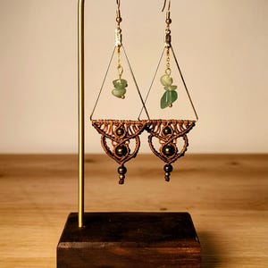 Könnte beinhalten: Ein Paar handgefertigte Ohrringe, präsentiert auf einem goldfarbenen Ständer. Die Ohrringe haben ein dreieckiges Design mit Makramee-Details, kleinen grünen Steinen und goldfarbenen Metallakzenten. Sie hängen an goldfarbenen Haken.