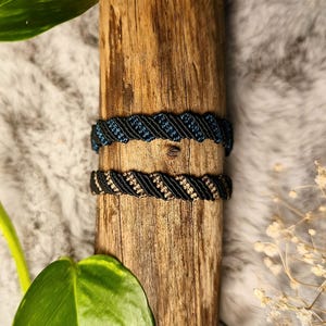 Peut inclure: Deux bracelets en macramé, l'un bleu marine et noir, l'autre noir et beige, sont présentés sur un morceau de bois vieilli. Les bracelets ont un motif tissé. L'arrière-plan comprend une texture douce et duveteuse et de la verdure.