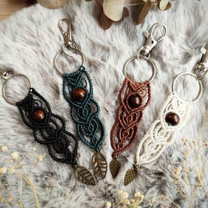 Peut inclure: Quatre porte-clés macramé noirs, turquoise, marron et blancs. Chaque porte-clés est orné d'une perle en bois et d'un charm en forme de feuille métallique. Présentés sur une surface grise et duveteuse.
