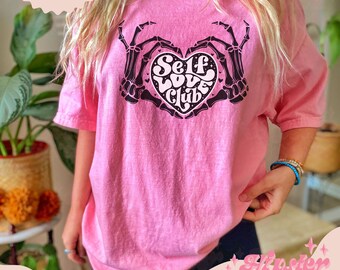 May include: Pink t-shirt with a black graphic of a heart with the text "Self Love Club" inside. The heart is held by two skeletal hands.