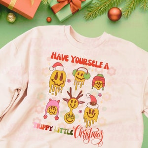 May include: A pink sweatshirt with a Christmas-themed design. The design features a smiling face with a Santa hat and the text "Have Yourself a Trippy Little Christmas".