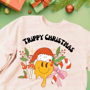 May include: A pink sweatshirt with a trippy Christmas design. The design features a melting smiley face wearing a Santa hat, mushrooms, candy canes, and a pink bow.