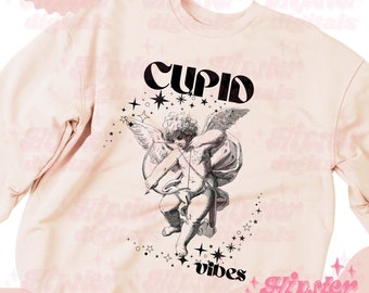 May include: A pink sweatshirt with a black graphic design of a cupid with wings and an arrow, surrounded by stars. The text "Cupid vibes" is below the cupid. The text "Hipster digitals" is in the bottom right corner.