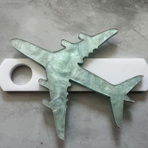 May include: A light green airplane shaped paperweight with a glittery finish. The paperweight is sitting on a white rectangular base with a circular hole in the centre.