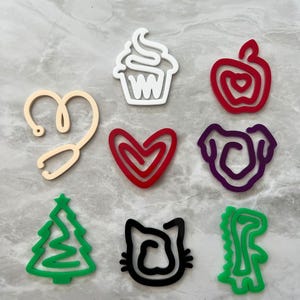 Puede incluir: Nueve formas de contorno de diferentes colores, incluyendo una magdalena, una manzana, un corazón, un perro, un árbol de Navidad, un gato y un dinosaurio. Las formas están cortadas en acrílico y tienen aproximadamente 5 cm de tamaño.