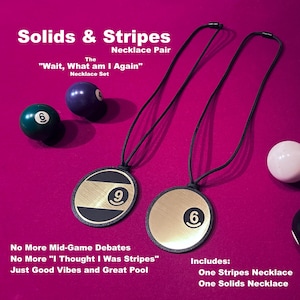 May include: A necklace pair with black cords and circular pendants. One pendant features a gold-colored face with a black stripe and the number 9. The other pendant has a gold face with the number 6. The text reads "Solids & Stripes Necklace Pair".