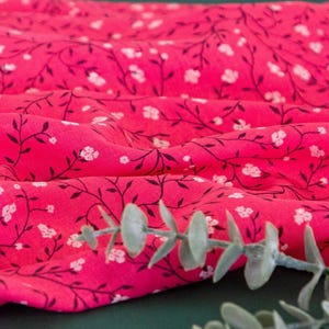 Może przedstawiać: Różowy materiał z białym wzorem kwiatowym. Materiał jest udrapowany na zielonej powierzchni z gałązką zielonych liści.