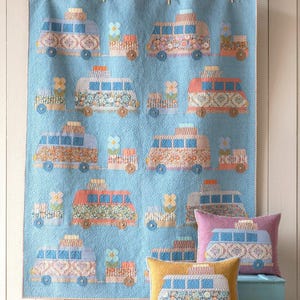 May include: A light blue quilt with repeating patterns of colorful camper vans and flower accents. The quilt is displayed with three decorative pillows, each featuring a camper van design. The pillows are in shades of yellow and pink.