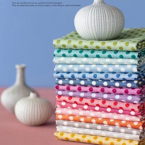 May include: A pile of multi-coloured polka dot fabric, with a white ceramic vase on top. The fabric includes shades of green, blue, pink, and orange. The text reads: "Dots are versatile and can be used for all kind of projects. They are especially sweet as lining in bags or as binding on quilts and pillows."