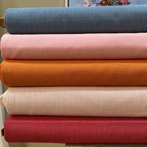 Op de afbeelding: Vijf rollen gestreepte stof in verschillende kleuren. De stof is op elkaar gestapeld. De kleuren zijn blauw, roze, oranje, wit met roze strepen en rood.