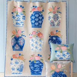 May include: A beige quilt and two pillows feature a repeating pattern of blue and white vases filled with pink, orange, and blue flowers. The quilt is hung on a light blue wall, and the pillows are on a wooden surface.
