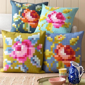May include: Four decorative throw pillows with floral designs. The pillows feature a pixelated floral pattern in shades of pink, orange, blue, and yellow. The pillows are arranged on a beige sofa, with a tray and decorative items in the foreground.