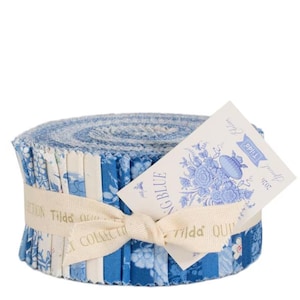 May include: A roll of blue and white fabric strips, tied with a cream ribbon. The fabric features floral and solid patterns. A tag with floral illustrations and the text "Spring Blue" is attached to the roll. The ribbon is printed with the text "Tilda Collection".