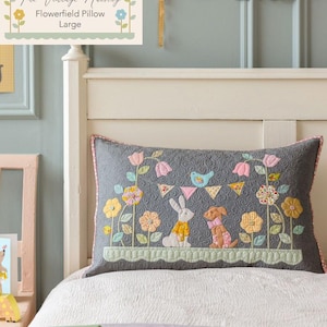 May include: A large, gray decorative pillow with a floral and animal design. The pillow features a bunny and dog, surrounded by flowers and a banner. The pillow is on a bed with a white headboard. The text on the pillow reads "Flowerfield Pillow Large".