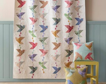 May include: A white quilt with a pattern of colorful birds. The quilt is hanging on a clothesline and there are two pillows on a yellow stool in front of it.