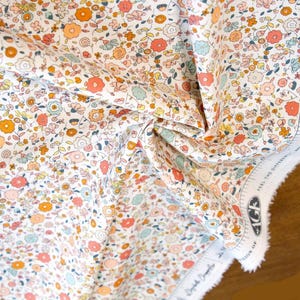 May include: White cotton fabric with a small-scale floral print in shades of orange, pink, and blue. The fabric has a white selvage edge with text that reads "100% Premium Cotton, Fiber Junctions.com, Oeko-Tex Standard 100."