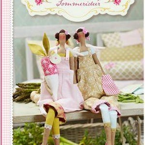 Puede incluir: Dos muñecas hechas a mano con diademas florales y delantales. Una muñeca sostiene un objeto con forma de zanahoria. Las muñecas están sentadas en una mesa con espárragos y otras verduras. La imagen tiene una paleta de colores suaves.