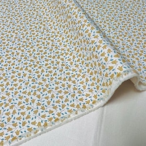 Op de afbeelding: Witte stof met een herhalend patroon van kleine gele bloemen met blauwe stelen. De stof is dubbelgevouwen en de rand is zichtbaar.