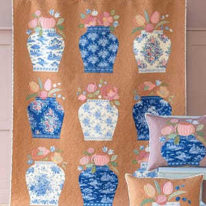 May include: A quilted wall hanging and two throw pillows featuring floral arrangements in blue and white vases. The vases are filled with pink and peach flowers. The background is a textured, warm brown color.