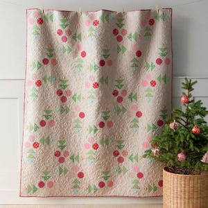May include: A cream-colored quilt with a red, pink, and green geometric pattern. The quilt is hanging on a white wall with a small Christmas tree in a wicker basket in the foreground.