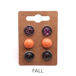 May include: Three pairs of stud earrings mounted on a rectangular brown card. The top pair features purple glitter, the middle pair is solid orange, and the bottom pair is black with multicoloured flecks. The word FALL is printed at the bottom.