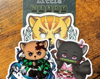 May include: Three colorful stickers featuring anime characters with cat ears. The stickers are on a wooden surface. The characters are a yellow cat with a white face, a brown cat with a green and black outfit, and a black cat with a pink and red outfit.