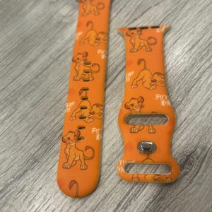 May include: Orange watch band featuring Simba from The Lion King. The band has a repeating pattern of Simba in various poses. The text "Future King" is also printed on the band. The band is made of a flexible material.