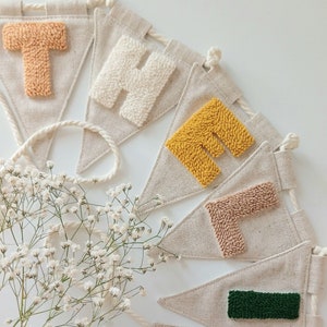 Puede incluir: Un banner con la palabra "HELLO" escrita en letras de hilo de colores. Cada letra está en una bandera triangular separada hecha de tela de lino natural. Las banderas están conectadas por una cuerda blanca.