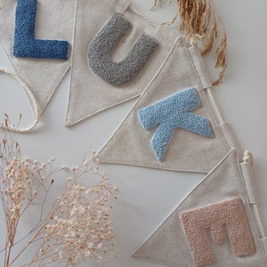 May include: A banner with the name "LUKE" spelled out in colorful yarn letters. Each letter is on a separate triangular flag made of natural linen fabric. The flags are connected by a white string.