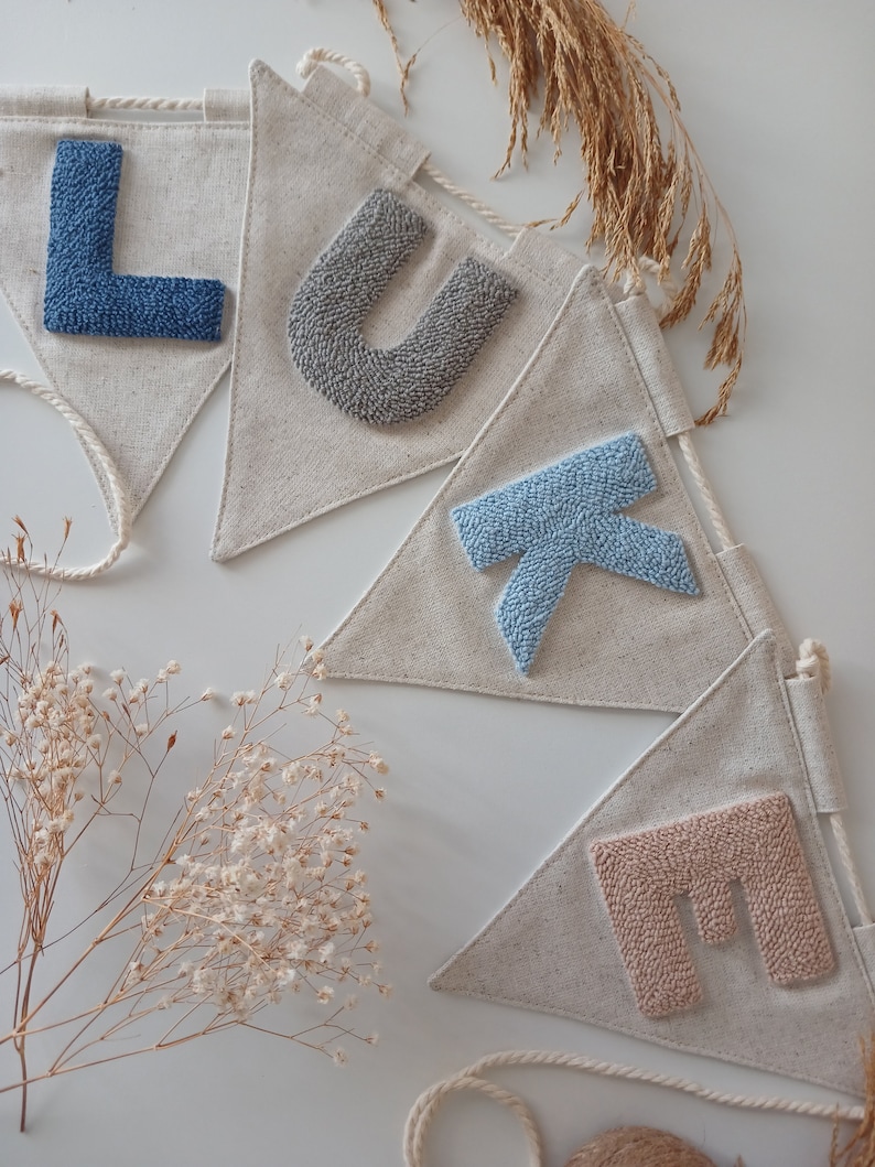 May include: A banner with the name "LUKE" spelled out in colorful yarn letters. Each letter is on a separate triangular flag made of natural linen fabric. The flags are connected by a white string.