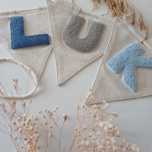 May include: A banner with the letters "LUK" spelled out in blue and gray yarn. The letters are attached to a white fabric banner with a rope attached to the top.
