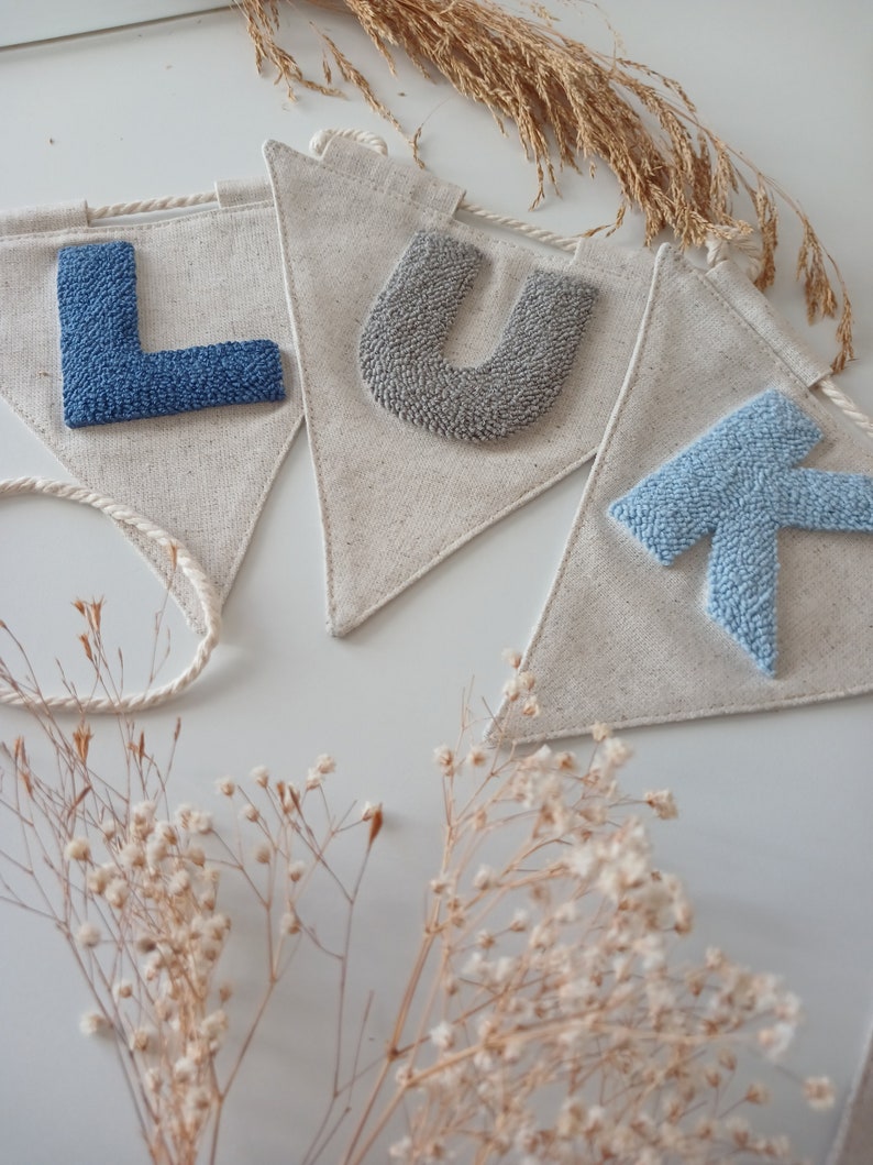 May include: A banner with the letters "LUK" spelled out in blue and gray yarn. The letters are attached to a white fabric banner with a rope attached to the top.